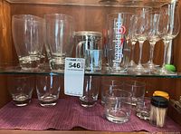 Glass cabinet shelf with assorted drinking glasses including beer glass, water glasses, toothpick holder with toothpicks, and short glasses.