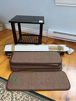 Photo showing one dark brown side table, a long boxed item (grab bars) and stacked brown stair protector mats on wooden floor.