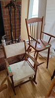 Two wooden chairs, one a child size rocking chair with vinyl seat/back and another vintage wood doll chair without tray, placed near a window on wooden floor.