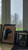 Photo showing multiple framed prints of black and golden Labradors stacked on floor near window, varying sizes.