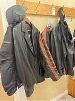 Lot of four leather jackets hanging on coat hooks alongside matching leather chaps; includes a black leather blazer, black and brown striped motorcycle jacket, plain black leather jacket, textile motorcycle jacket, and leather chaps.