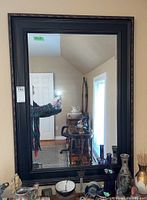 Full view of the large rectangular wall mirror with black carved frame hanging on a wall. Reflections of room interior and door visible.
