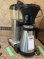 Front angle view showing stainless steel and black plastic coffee maker with measuring water reservoir, thermal carafe, and base with power switch.