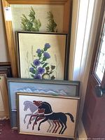 Four framed embroidered pieces leaning against a wall; pine tree embroidery at the back, purple thistles embroidery next, with two framed needlepoint horses in front, all on a carpeted floor.