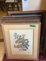 Group of framed needle art pieces stacked together showing multiple frames including one with cross stitch of birds and holly with wear on edges.