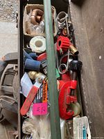 Close-up photo of plumbing repair tools and fittings including copper elbows, pipe cutter, pipe wrench, valves, and plumbing tape in a metal toolbox.