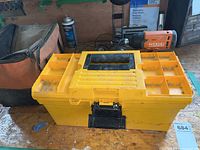 Yellow plastic toolbox closed on a wooden surface with one Ridgid cordless power drill standing upright beside it. An orange-black tool bag is visible to the left.