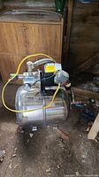 Home Plumber Professional Series HPS-75SS water pump with stainless steel tank and motor assembly, yellow power cord visible, located in a wooden shed on a dirt floor.