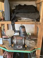 Full view of Ryobi bench grinder showing grinding wheels and attached light, with various tool belts and bags above it on the shelf.