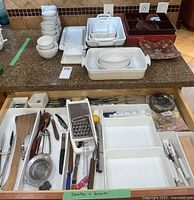 Drawer with assorted kitchen tools including spoons, tongs, graters and utensils, along with additional serving ware dishes on counter above.