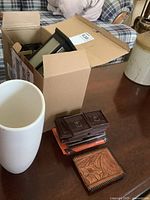 Box of eight black outdoor solar mountable lamps with clear front panels and black bases shown on a wooden table next to a tall white ceramic vase, several rubber floor protectors, and a tooled leather wallet.