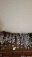 Wide view of large collection of glass stemware on wooden surface showing multiple rows of wine glasses and champagne flutes.