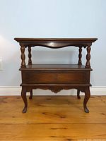 Full front view of vintage style wooden side/end table showing upper shelf, turned legs, lower drawer and curved legs.