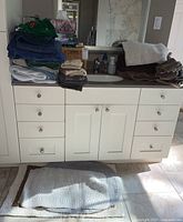 Stacked bath towels of various colors and textures on bathroom counter with wicker basket and tissue box behind, plus bath mats on floor in front.