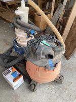 Vacuum with cyclone dust collector system and connected hoses placed on concrete floor in a workshop area, power cord coiled on top.
