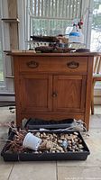 Photo of vintage wood cabinet with dovetailed drawer, metal pulls, and double doors; includes tray with decorative stones and miscellaneous items below cabinet.