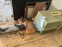 Closed and partially open green tackle box with a smaller translucent orange lure container and a few loose lures and soft plastics on a wooden surface