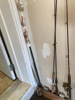 Photo showing three fishing rods leaning against a wall, two with reels attached, cork handles visible. Set of spinner fishing lures hanging vertically beside rods.