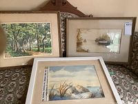 Three framed watercolors arranged on patterned sofa: forest scene on left, river boat on right, beach scene foreground.