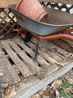 Side view of metal wheelbarrow with one rubber tire wheel, orange frame, and blue basin containing terracotta pot.