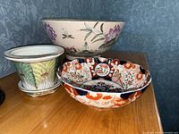 Decorative bowls and planter shown together on wood surface. Bowls have floral and geometric patterns in multiple colors. Planter has leaf design in green with brown.
