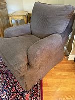 Side angle view of one of the large brown corded fabric armchairs showing the cushioned seat, back, and armrest along with the skirted base.