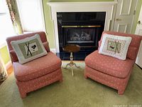 Two coral boudoir chairs with floral upholstery and sushi needlepoint pillows. Small round hand-painted table between them in front of fireplace.
