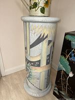 Tall cylindrical plant/art stand with crackled paint finish and abstract design, showing floral and geometric patterns, placed near a corner and next to rectangular stand.