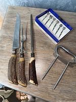 Meat carving knife, carving fork, sharpening steel with antler handles, sterling silver cocktail picks in blue box, and metal meat spike on wood surface.