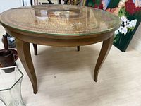Full view of round wooden coffee table with curved legs and glass top over embossed leather surface.