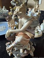 Porcelain figurine of seated woman with angel on the left, showing hand-painted floral dress details and some damage.