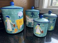 Four hand painted Susan Sargent ceramic canisters of varying sizes with lids, painted with pastoral farm animal and woman designs on turquoise background.