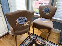 Two upholstered chairs side by side by window, showing dark beige fabric with blue floral pattern, wooden legs, and decorative nailhead trim.