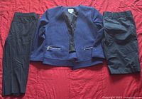 Blue blazer with textured weave and zippered pockets laid flat on a red surface, shown with two pairs of black pants folded beside it.