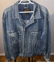 Front view of large vintage Eddie Bauer denim jacket hanging on white plastic hanger, showing button closure and two chest pockets with button flaps.