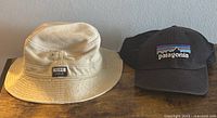 Two hats shown on wooden surface: Nike Golf beige bucket hat and black Patagonia snapback hat with mountain logo.