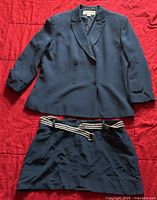 Blazer and mini skirt laid flat on red fabric background, showing front view with double breasted navy blazer and skirt with striped belt.