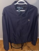 Front view of navy blue Polo Ralph Lauren jacket hanging on a hanger on a chair, showing full zipper closure and embroidered Polo logo on left chest.