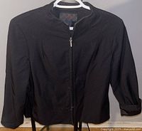 Front view of black nylon women's jacket hanging on white plastic hanger showing collarless neckline, long sleeves, front zipper and drawstrings at bottom hem.