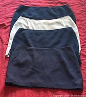 Four mini skirts laid flat, showing front side, in dark navy, medium navy, light khaki, and dark blue hues against a red background.