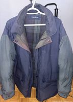 Front view of the navy blue and gray men's Nautica jacket hanging on a white hanger, showing snap buttons, zippers, and plaid lining inside.
