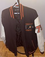 Front view of the black varsity jacket with white sleeves showing Philadelphia Flyers logo and 'Stanley Cup Champion' text on chest, and '74' on sleeve.