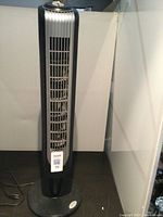 Front view of the black oscillating tower air fan, tall with vertical slats and power cord visible on the floor.