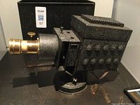 Side view of the antique picture projection unit showing textured black metal body, brass lens, mounting base and adjustment knob.