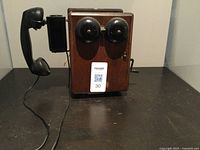 Front view of antique wooden wall phone with dual bells, crank, and wired handset.