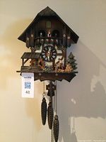 Front view of wooden chalet-style cuckoo clock with pinecone weights hanging against beige wall