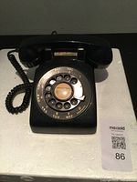 Front view of vintage black rotary dial telephone with dial numbered 0-9 and corresponding letters, coiled cord connected to the handset; placed on a white surface.