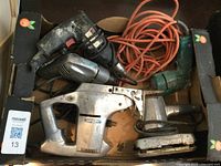 Photo showing six vintage electrical hand tools including different types of drills, jig saw, sander, and orange extension cord placed in a cardboard box.