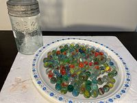 Photo showing approximately 90 colorful glass marbles on a white plate with blue floral border alongside a vintage Crow glass jar with metal lid on a dark surface.