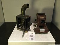 Photo shows two vintage projection devices side by side on a white surface: a metal Magic Lantern with chimney pipe and a brown picture projection unit.